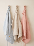 Three textured cotton muslin cloths in light blue, cream, and dusty pink, hanging from wooden pegs on a warm beige wall.