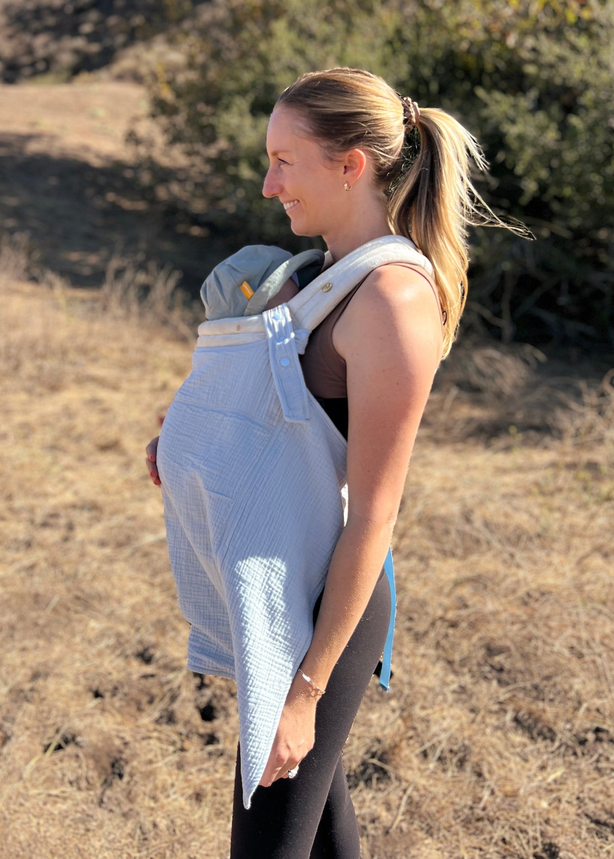 A woman wearing a baby in a carrier with a light blue sunshell cover outdoors in a dry grassy field.